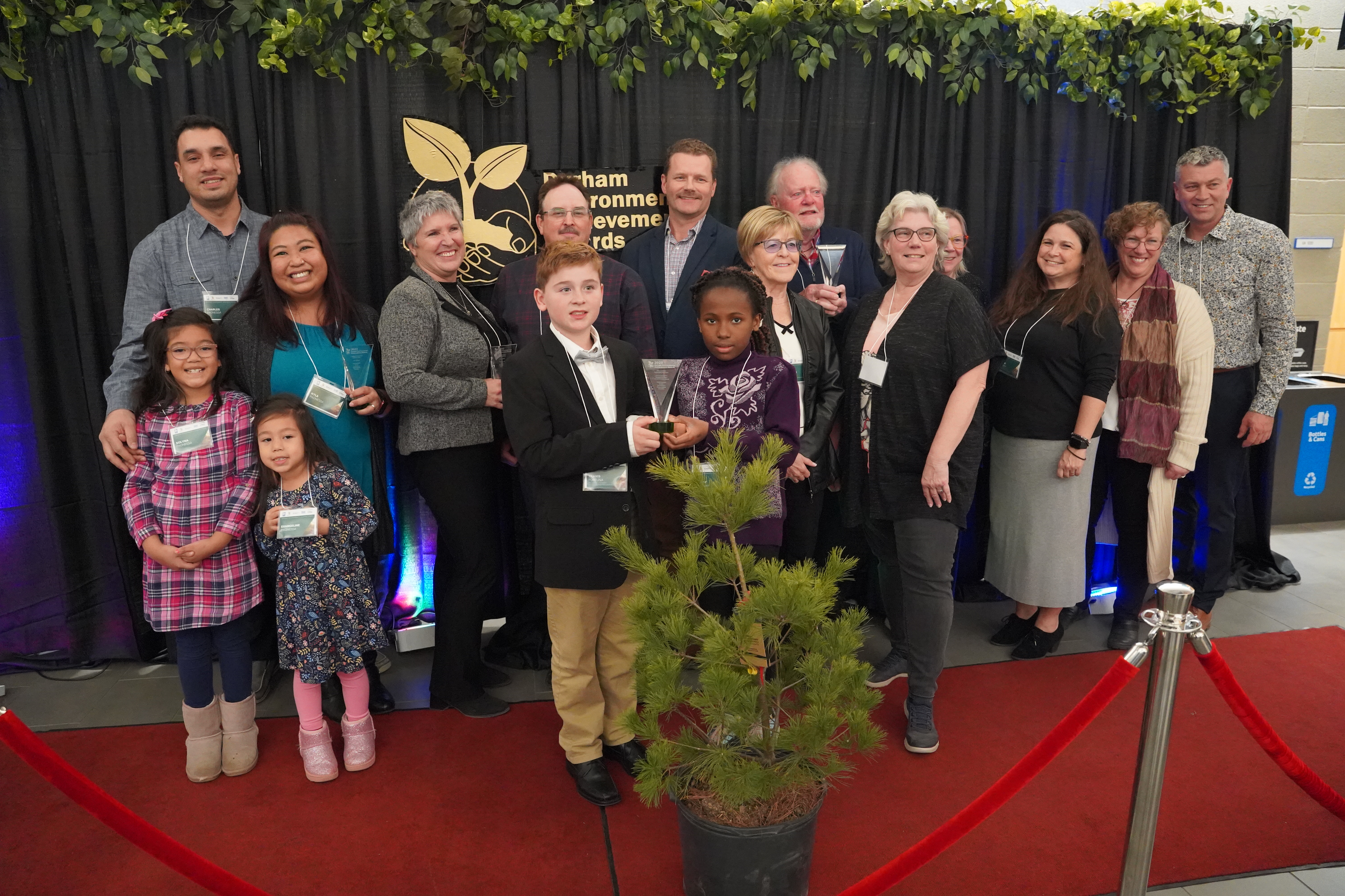 A large group of award winners, of all ages, stand on a red carpet with a black curtain behind them and a pine tree and red rope in front of them.