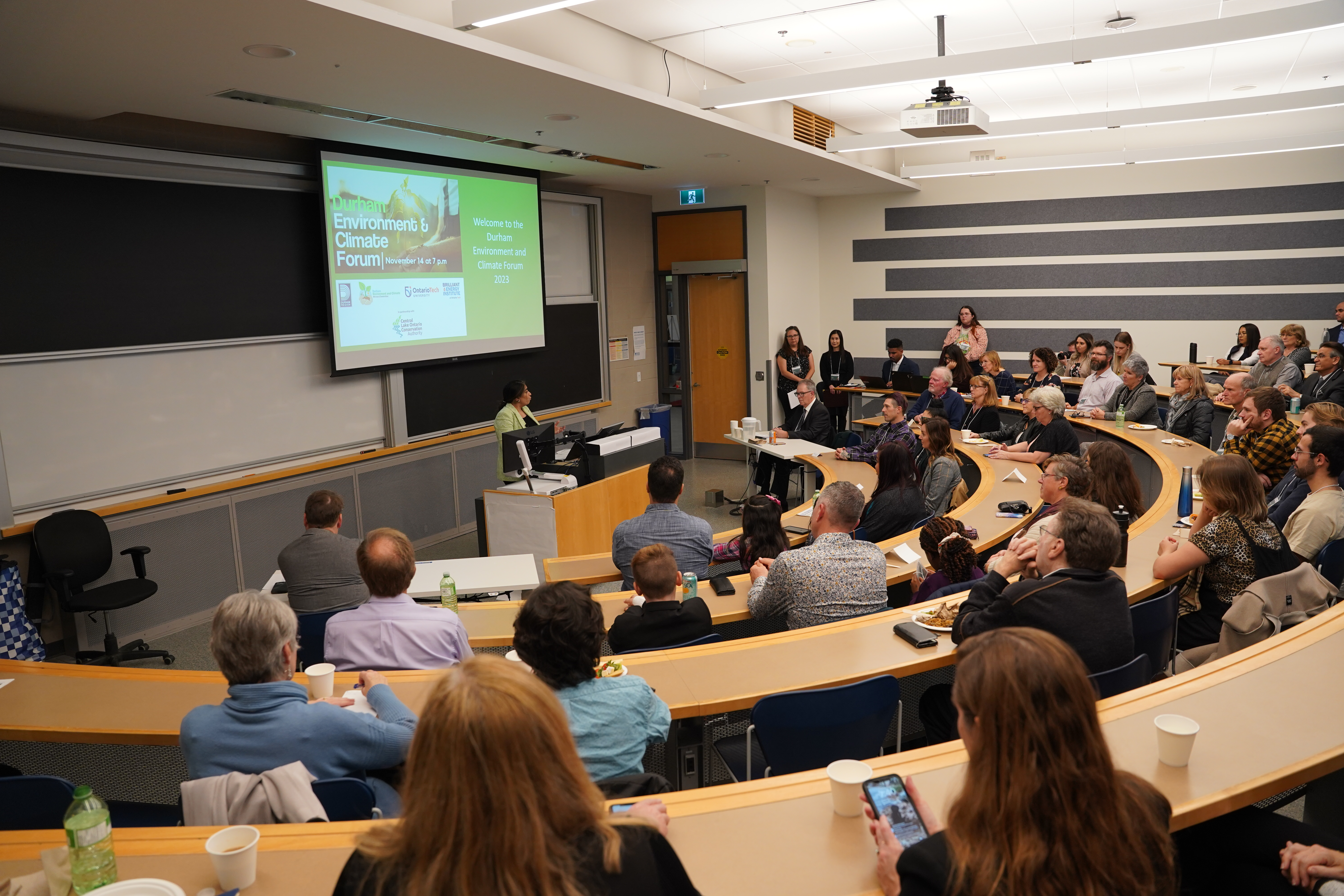 A woman stands at a podium with a presentation behind her in a full lecture room of people of all ages.